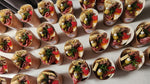 Load image into Gallery viewer, Multiple small paper cups arranged in rows on a white surface. Each cup contains sliced meats, olives, cheese, pickles, and garnish.
