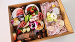 Load image into Gallery viewer, A gourmet gift box filled with an assortment of food items, chocolates, pamper items and flowers arranged in pink shredded paper inside a brown cardboard box.
